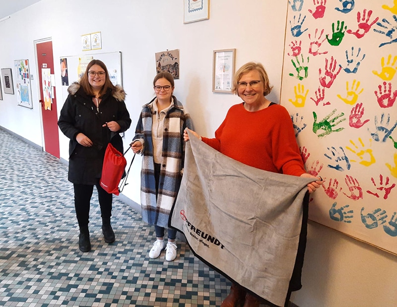 Drei Personen posieren mit einem PFREUNDT-Flaggenbanner neben farbenfrohen Handabdrücken an einer Wand.