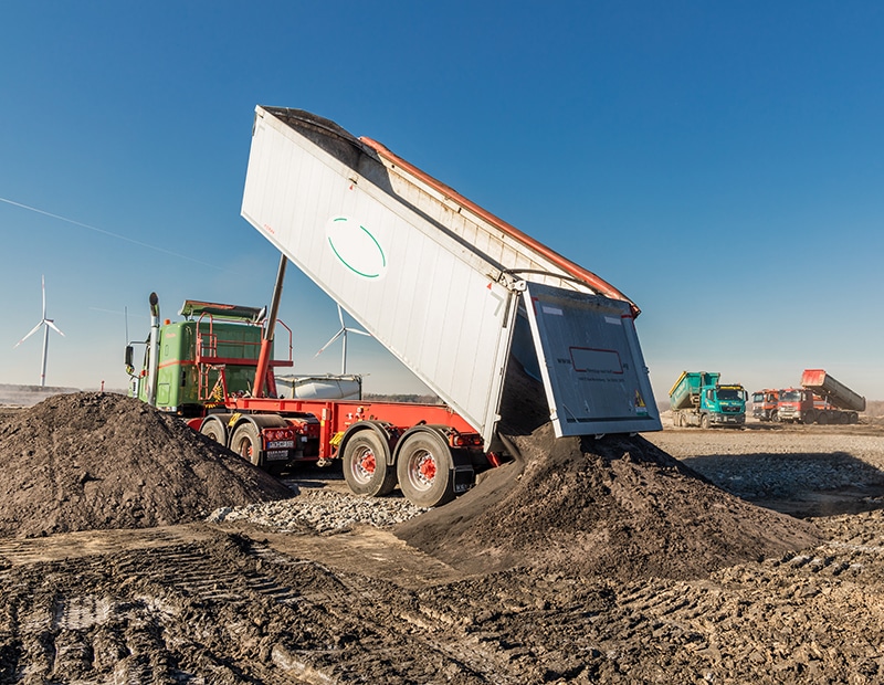 Truck unloading soil on a construction site under a blue sky, wind turbines in the background.