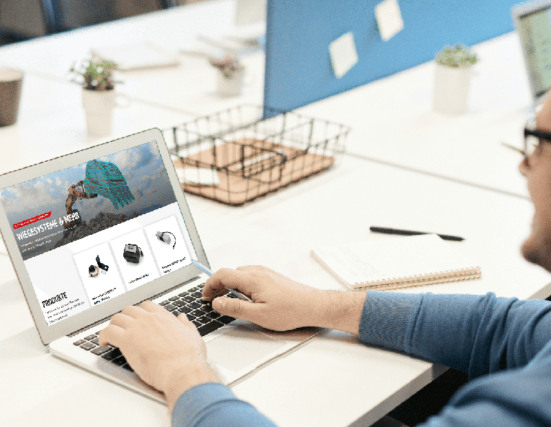 Person on a laptop viewing the PFREUNDT website for mobile weighing systems, including excavator, belt, and wheel loader scales.