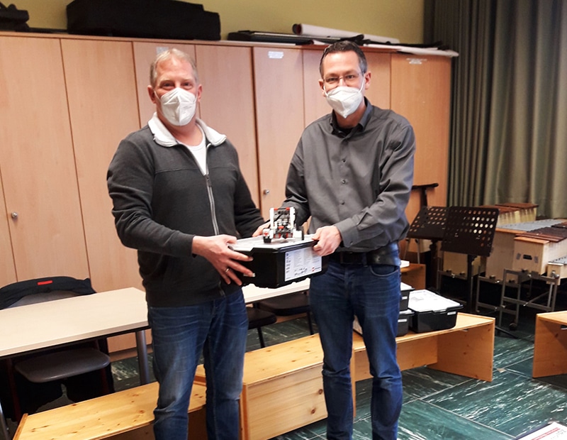 Zwei Männer mit Masken bei der Übergabe eines Lego-Roboters in einem Besprechungsraum.