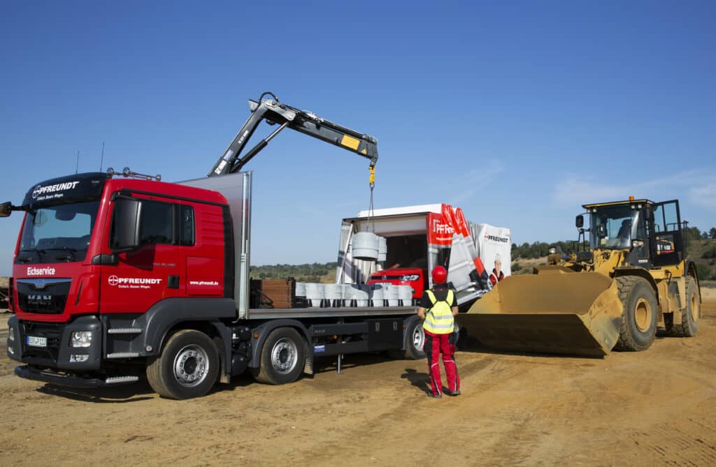 Roter PFREUNDT Eich-LKW mit Ladekran auf einer Baustelle, daneben ein Radlader und ein Mitarbeiter in Schutzkleidung.