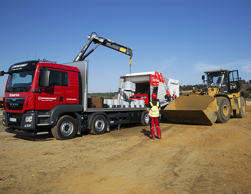 Red PFREUNDT calibration truck with loading crane on a construction site, next to a wheel loader and an employee in protective clothing.