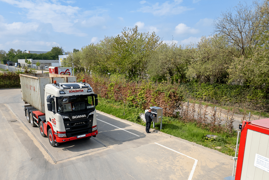 Truck on a PFREUNDT scale with operator at the control station, surrounded by green landscape and blue sky.