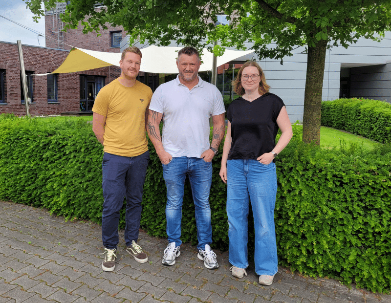 Three people smiling at the camera in front of a PFREUNDT company building, provider of mobile weighing systems.