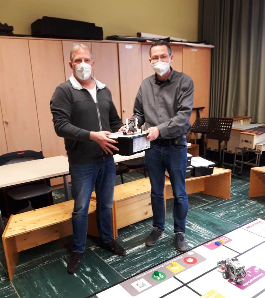 Zwei Männer mit Masken bei der Übergabe eines Lego-Roboters in einem Besprechungsraum.