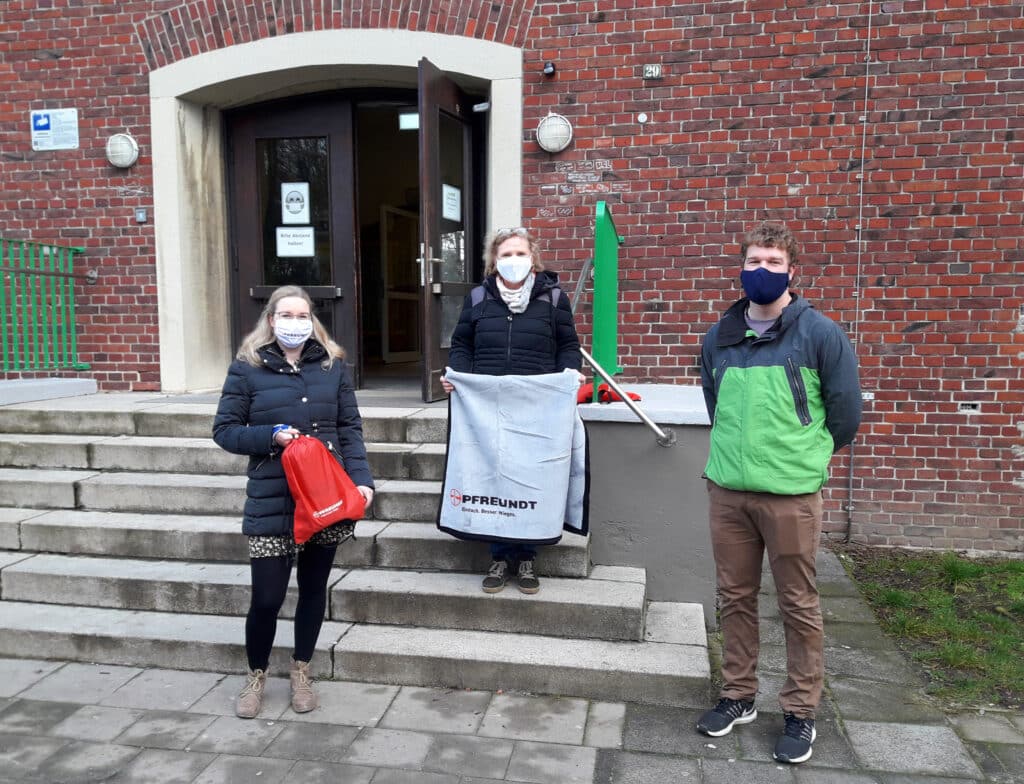 Personen vor einem Gebäude halten PFREUNDT-Produkte, tragen Masken und stehen auf einer Treppe.