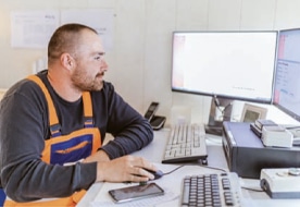 An employee in work clothes sits at a computer, analysing data for PFREUNDT’s mobile weighing systems, such as excavator and crane scales.