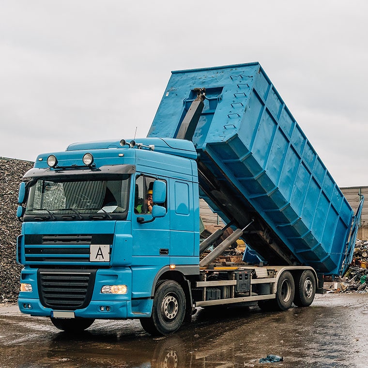Blue hook-lift truck with PFREUNDT Hook-Lift Scale tipping a container at a recycling yard.