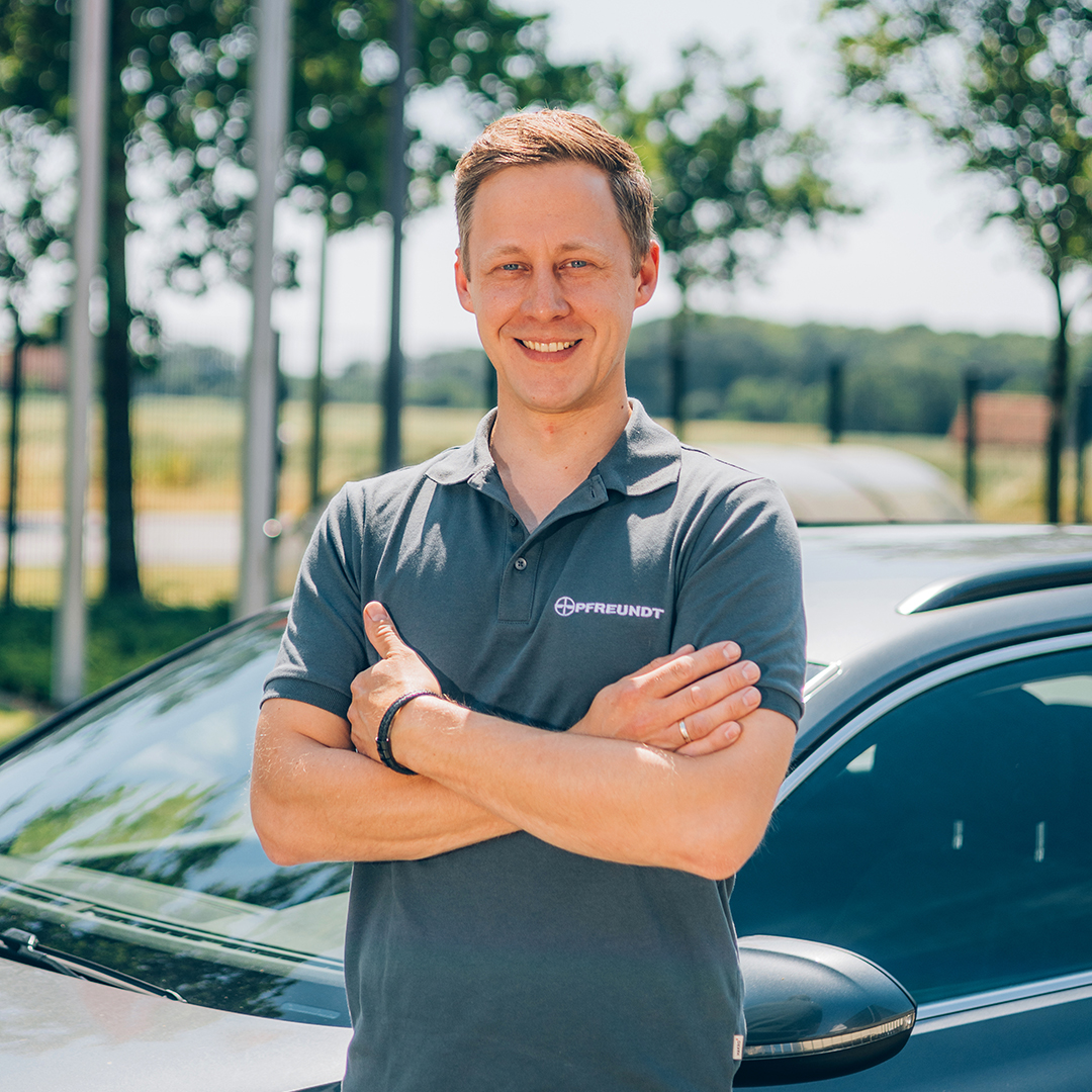 Person in a PFREUNDT polo shirt in front of a car, smiling with arms crossed, trees in the background.