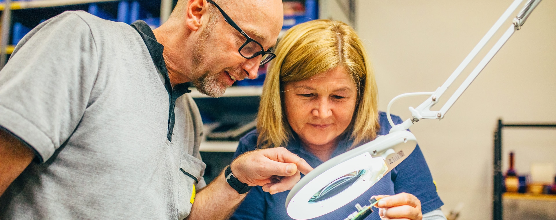 Dos personas trabajan concentradas en un componente electrónico bajo una lámpara de aumento en un laboratorio técnico.